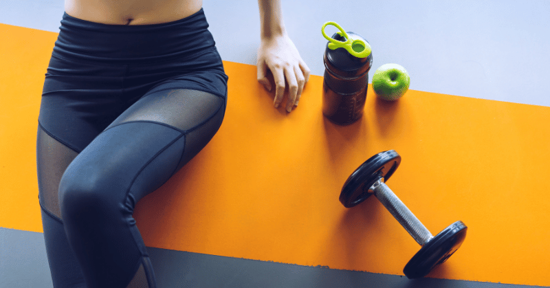 A woman's lower body on the gym floor with an apply, water bottle and dumbbell representing 30 day challenge ideas for physical fitness