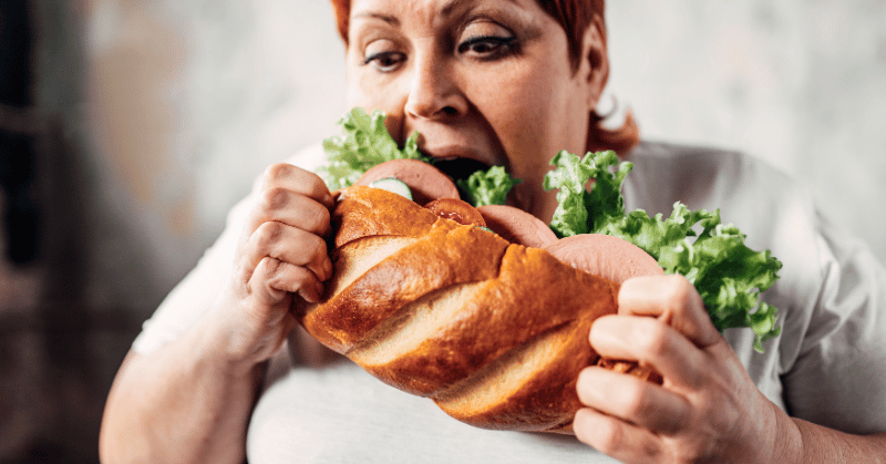 Overweight woman eating a large sandwich, demonstrating wasting time easting too much food