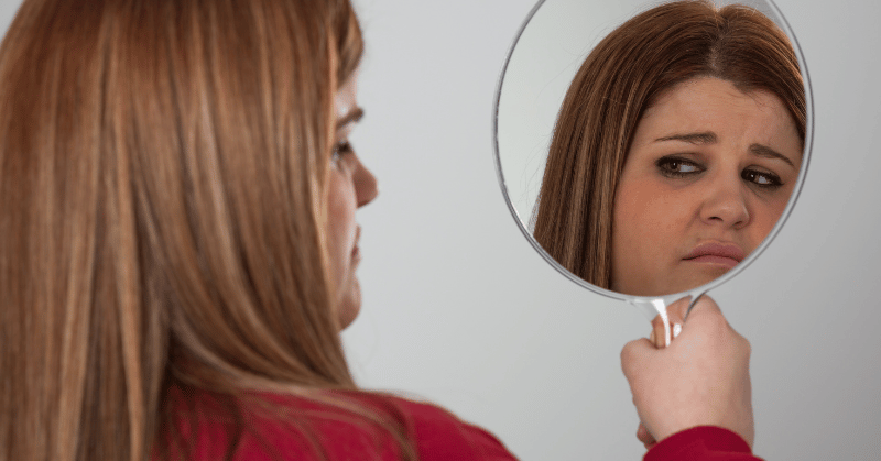 Woman looking herself in the mirror unhappily, displaying wasting time as a result of low self esteem