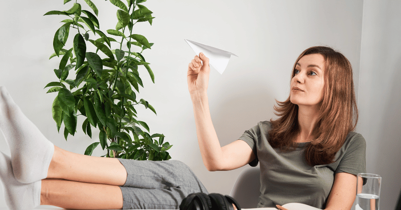 Woman procrastinating, sitting at desk and flying a paper plane, depicting wasting time